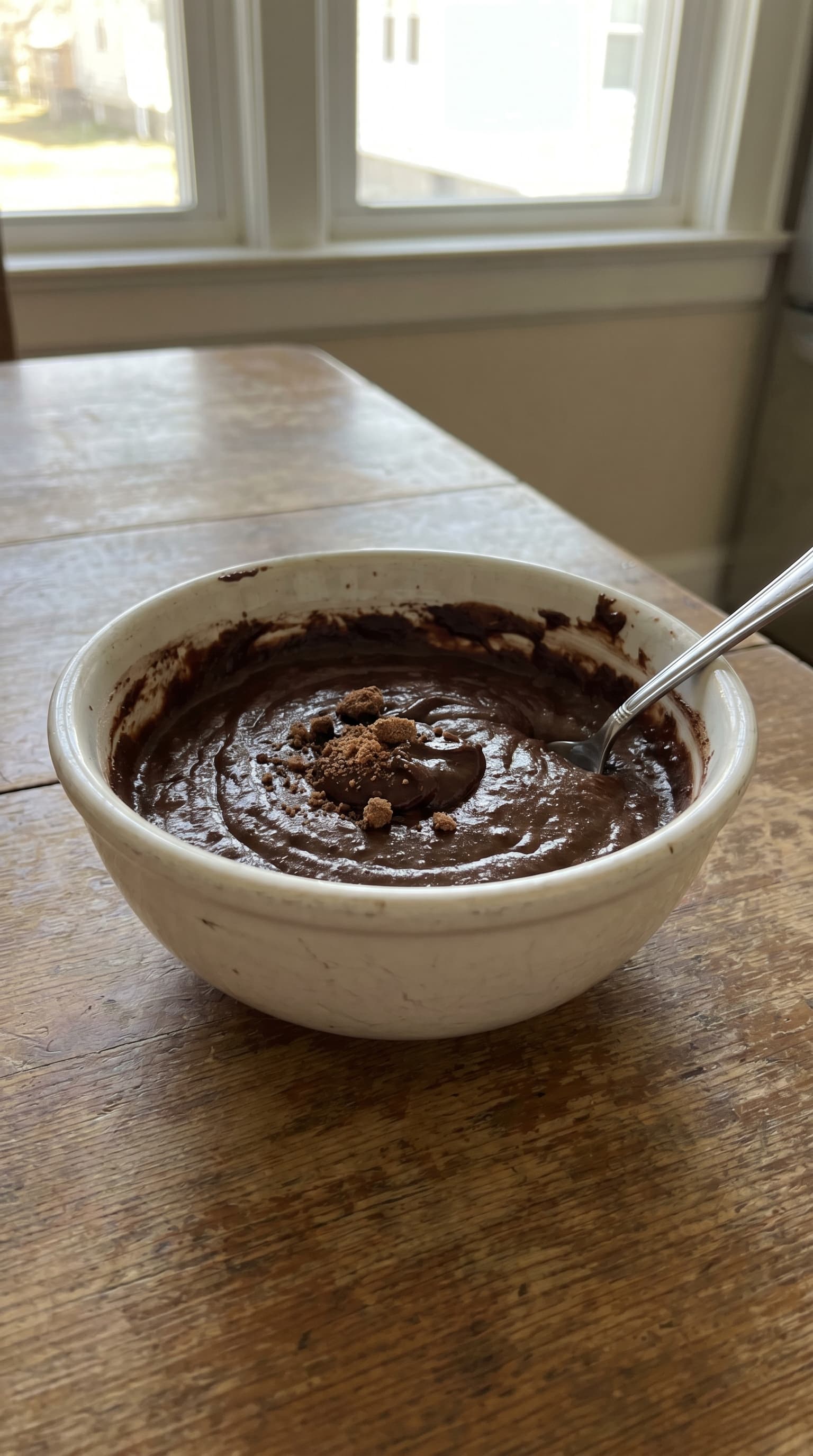 Thick chocolate protein brownie batter in a ceramic bowl with a spoon and cocoa crumbles on top by a kitchen window.