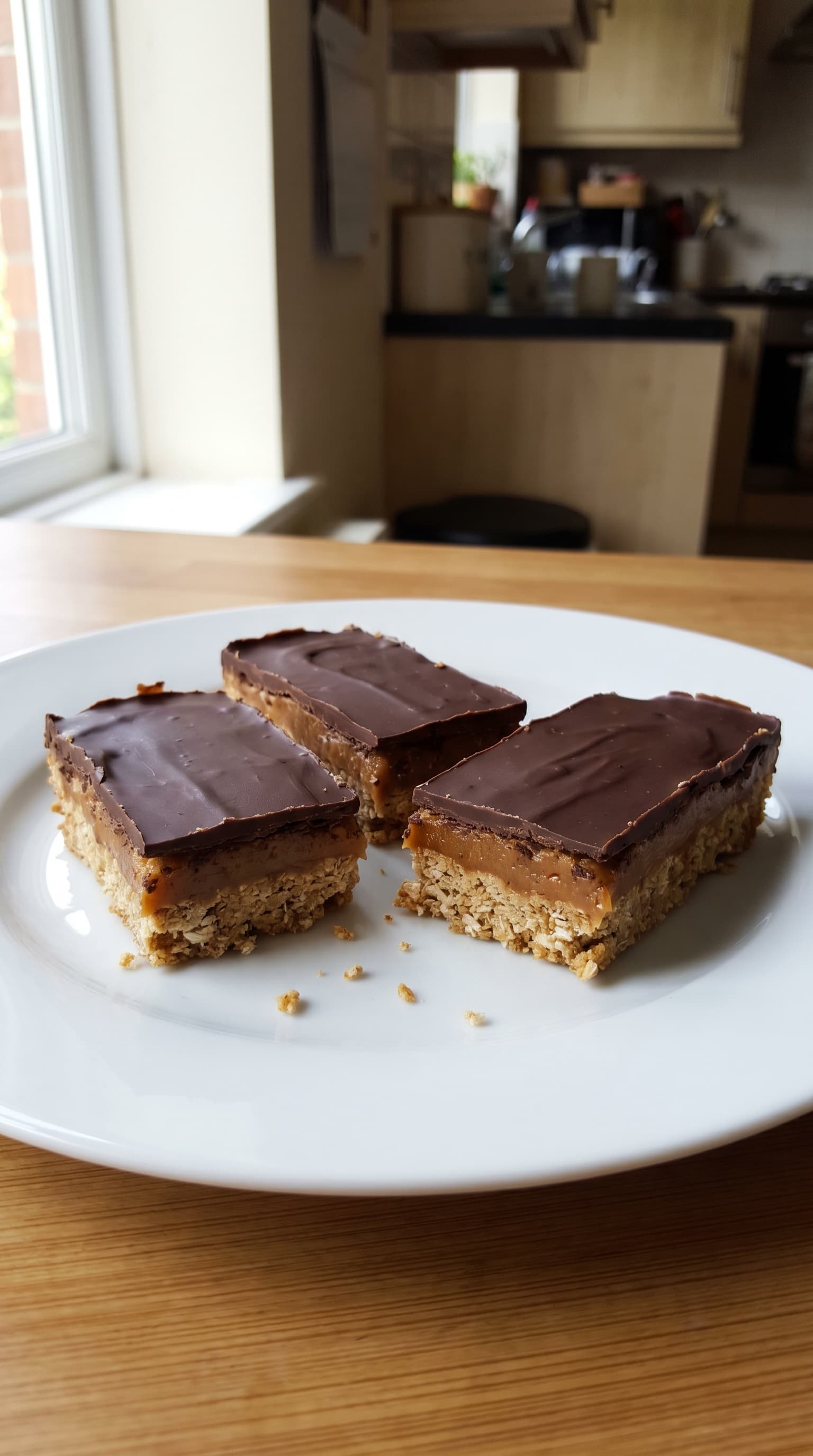 Twix protein bars with an oat base, caramel layer, and dark chocolate topping served on a white plate by a kitchen window.
