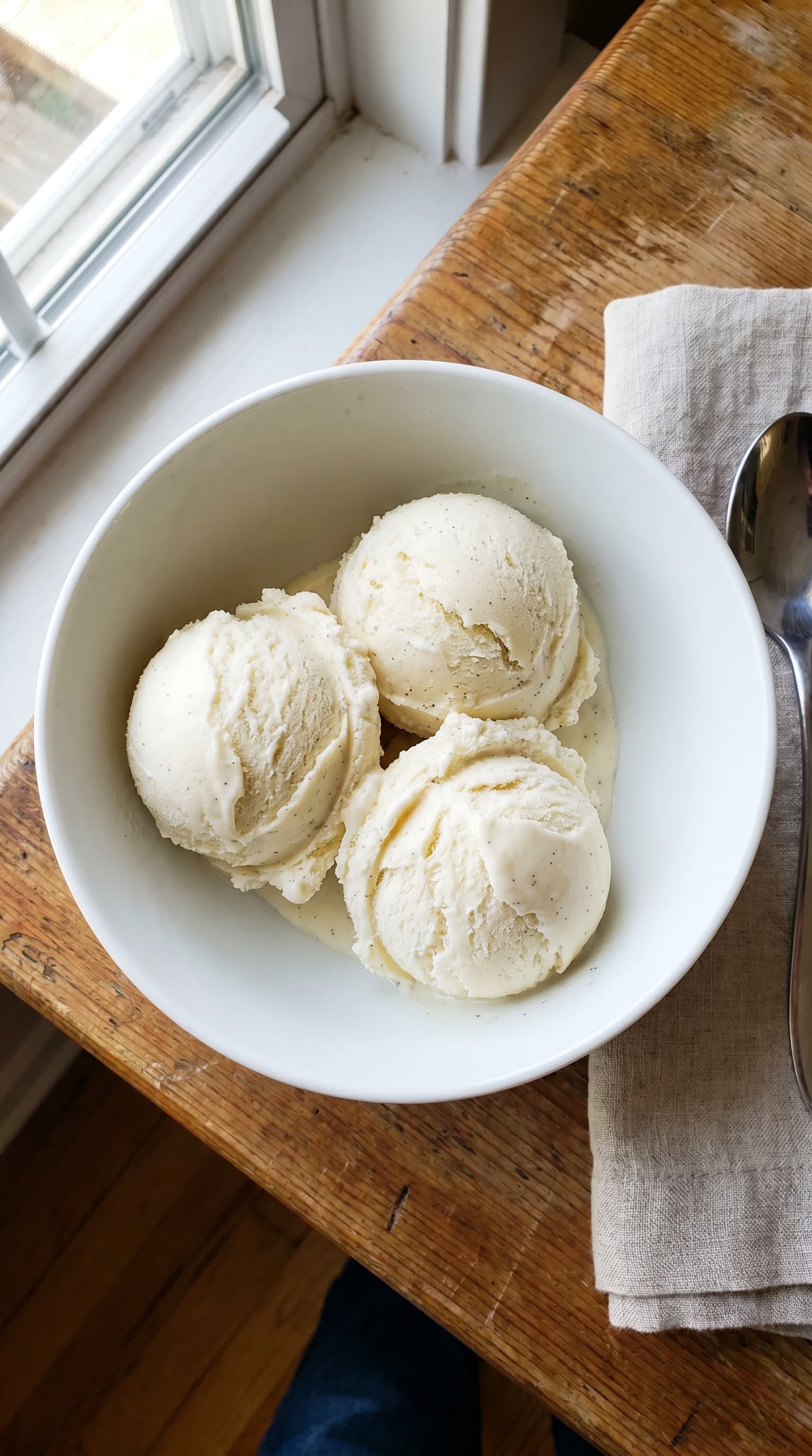 Three scoops of creamy vanilla ice cream in a white bowl beside a spoon on a wooden table by a window.